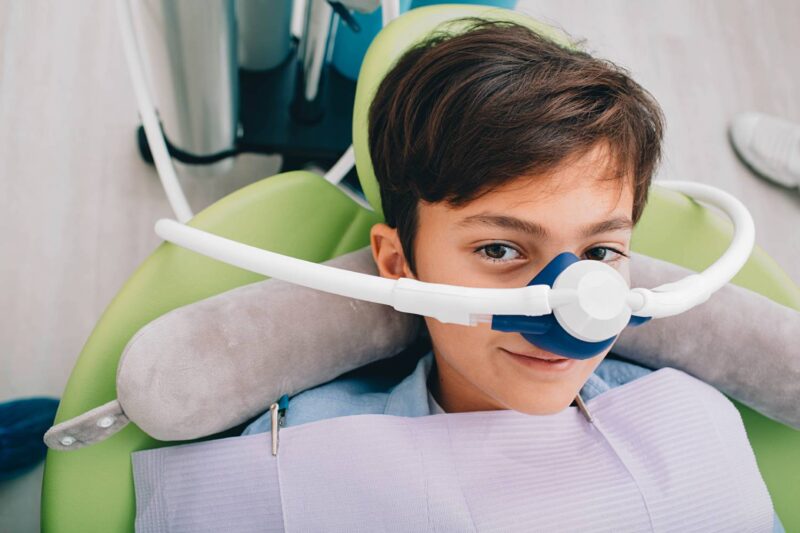 A child sits in a dental chair at Chestnut Dental Needham, wearing a nitrous oxide sedation mask and dental bib, preparing for a dental procedure.