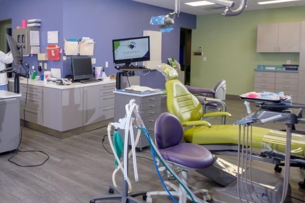 Modern dental clinic room at Chestnut Dental Needham with two dental chairs, medical equipment, cabinets, a computer, and a monitor displaying the clinic’s logo—serving Franklin, Bedford, and surrounding communities.