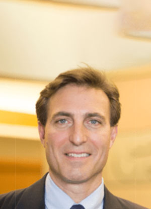 A man in a suit and tie is smiling at the camera with a blurred indoor background.