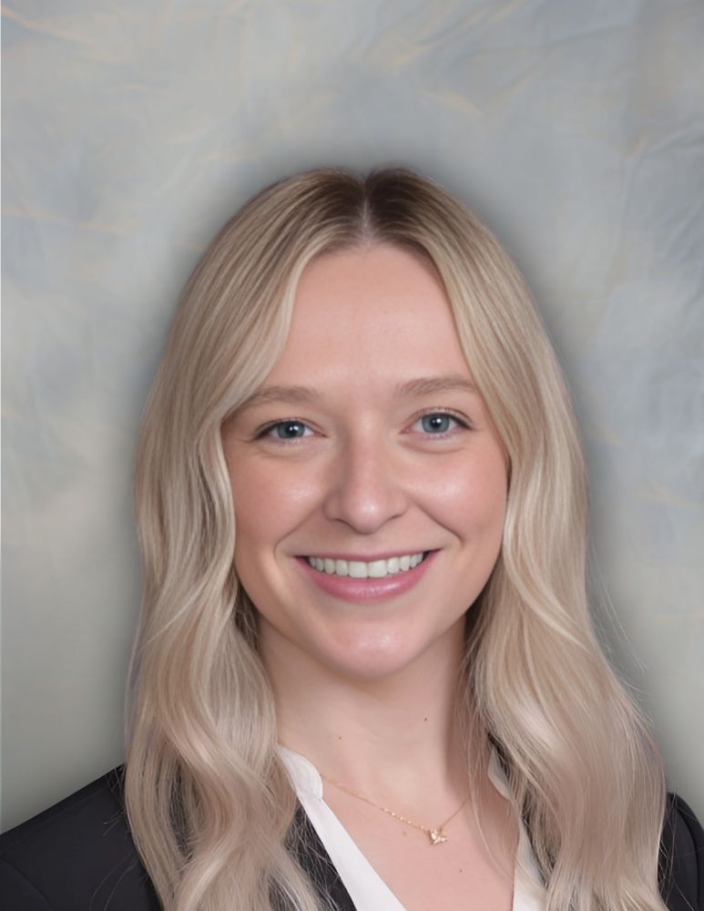 A woman with long blonde hair, wearing a black blazer and white blouse, smiles at the camera against a neutral, light-colored background.